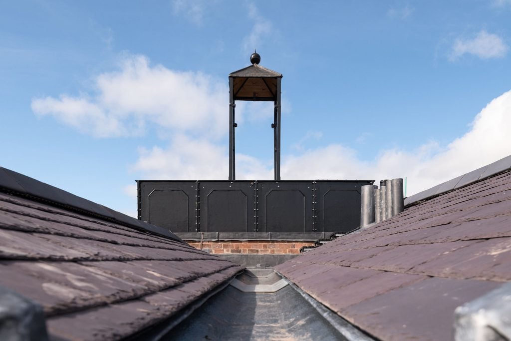 A photograph of a rooftop showing a large cast iron oblong water tank viewed from the side. There is a square structure with a pyramid roof on top of this. This structure once housed a bell.