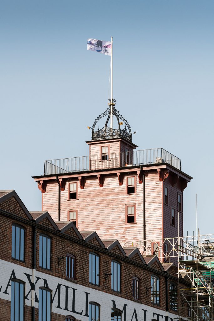 A photograph showing a flag flying above a three storey wooden tower with ornate ironwork coronet on top.